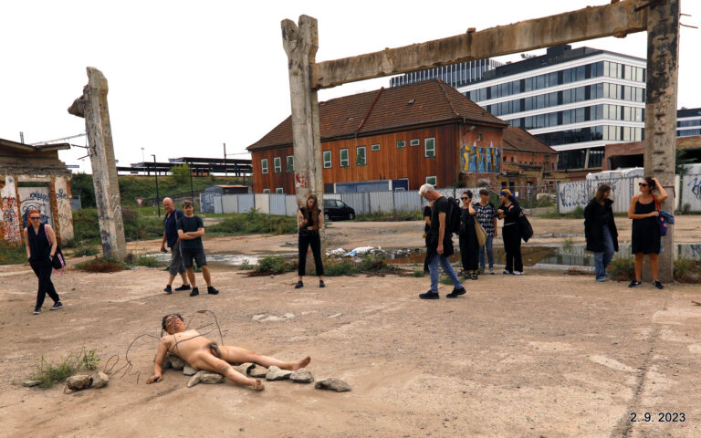 Een groep mensen in zwarte kleding kijkt naar een naakte performer die stenen op de grond rangschikt in een industrieel gebied met vervallen gebouwen op de achtergrond.