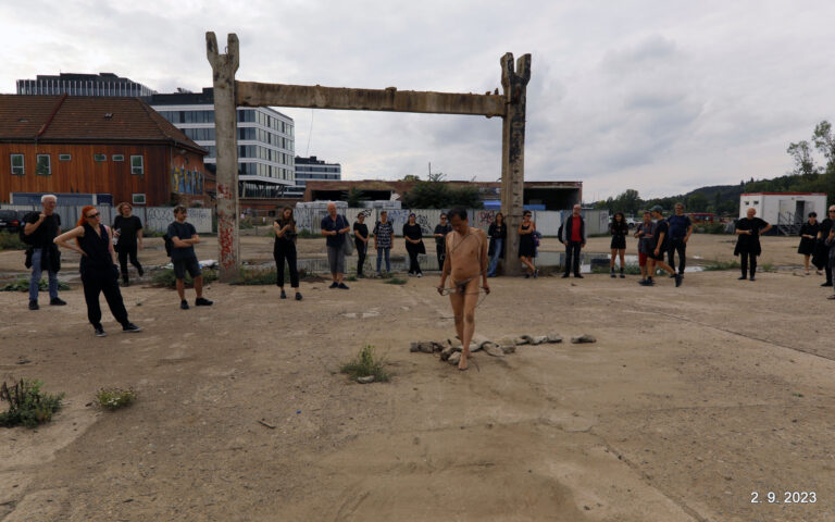 Een groep mensen in zwarte kleding kijkt naar een naakte performer die stenen op de grond rangschikt in een industrieel gebied met vervallen gebouwen op de achtergrond.