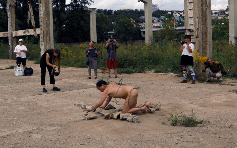 Een naakte performer is bezig met het plaatsen van stenen op de grond in een vervallen buitenomgeving, omringd door een groep toeschouwers die de actie fotograferen. Achtergrond met groen en fragmenten van afgebroken structuren.