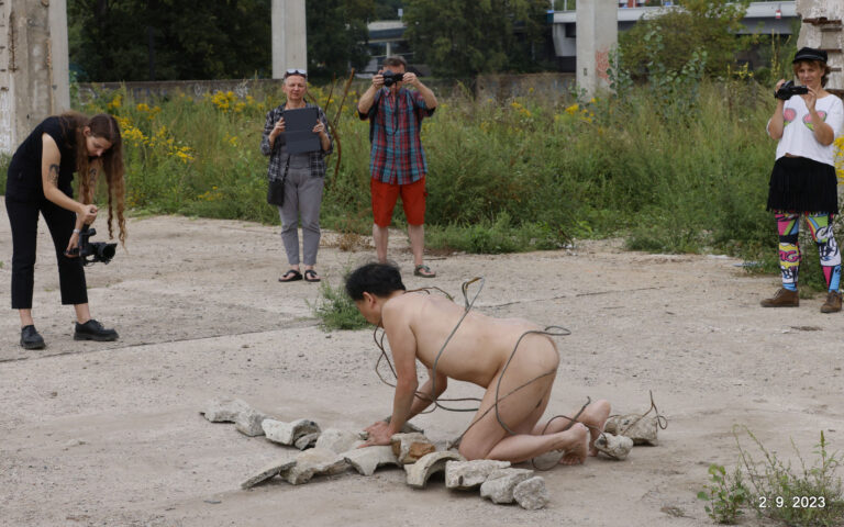 Een naakte performer, gebogen over een rij stenen terwijl hij omringd is door verschillende mensen die hem fotograferen. De omgeving is een ruwe, verwaarloosde buitenruimte met wilde bloemen en hoge grasplanten.