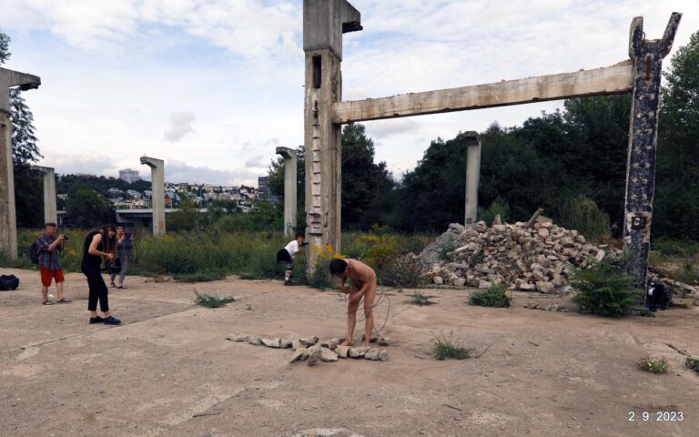 In een verlaten gebied, met ruïnes van betonnen constructies op de achtergrond, zijn verschillende mensen bezig. Een naakte persoon is vooraan bezig met een stapel stenen, terwijl achter hen enkele mensen met camera's foto's maken. Groene begroeiing groeit door de verwaarloosde omgeving.