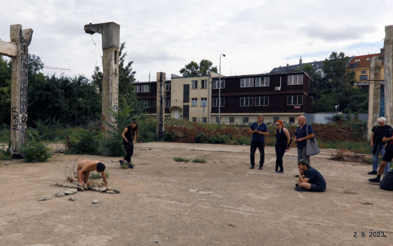 Een open terrein met afgebroken betonnen pilaren en begroeiing. In het midden knielt een naakte persoon bezig met stenen, terwijl een vrouw in zwart de situatie observeert. Groep mensen kijkt toe vanop een afstand, waaronder drie mannen en een vrouw. Op de achtergrond staan gebouwen en bomen onder een bewolkte hemel.