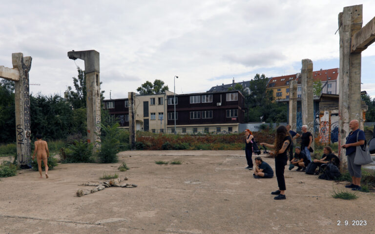 Een naakte persoon loopt door een vervallen, openluchtruimte met betonnen pilaren en ruïnes, omringd door mensen die toekijken. Op de achtergrond zijn appartementen te zien, met vegetatie en graffiti op de muren.