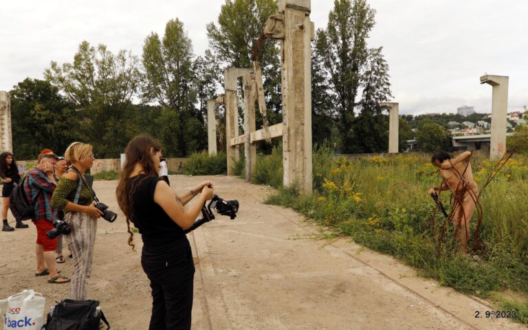 Een groep fotografen in een vervallen, groene omgeving met hoge struiken en bomen, terwijl een naakte model tussen takken poseert en een fotograaf hem vastlegt in de achtergrond.