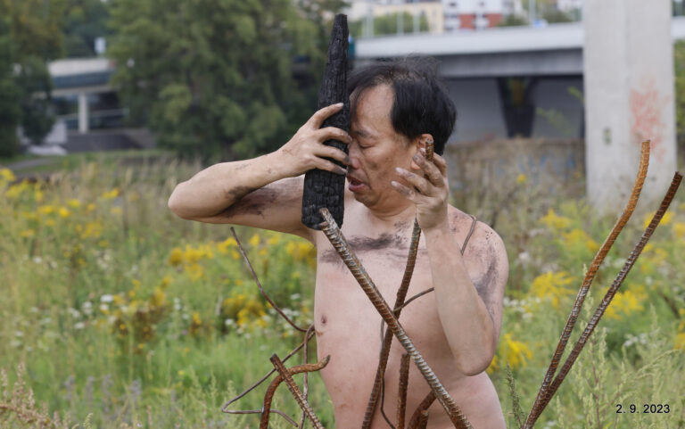 Een naakte man met zwartgemaakte huid staat in een veld vol gele bloemen. Hij houdt een zwart stuk hout tegen zijn voorhoofd en heeft verschillende metalen staven in zijn andere hand.