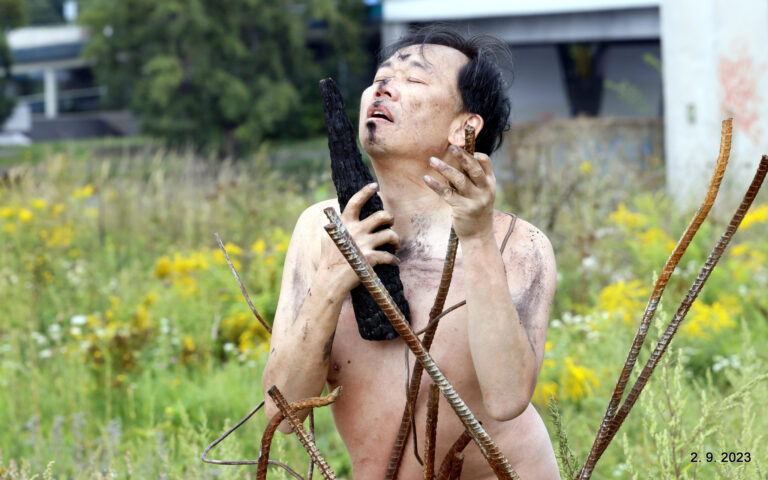 Een halfnaakte man met een bezorgde uitdrukking houdt een zwart houtachtig voorwerp vast, omringd door stalen staafjes, in een veld met bloemen en gras op de achtergrond.