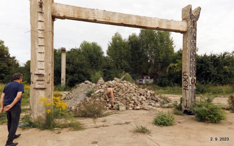 Een man met een blauwe t-shirt kijkt naar een grote hoop puin in een vervallen industriële omgeving, met bomen en planten op de achtergrond; op de hoop puin is een deel van een naakte persoon te zien die aan het graven is.