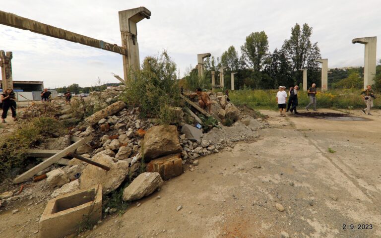 Een rommelige bouwplaats met stortstenen en een man in de schaduw van een berg puin, terwijl een groep mensen op de achtergrond wandelt tussen de afgebrande constructies en overgroeide gebieden op een bewolkte dag.