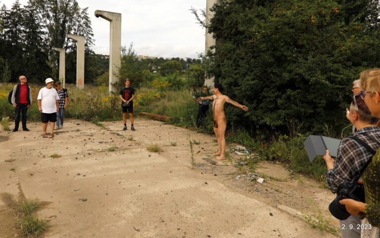 Een naakte man staat met uitgestrekte armen in een ruige, groene omgeving met achter hem enkele mensen die hem observeren. De achtergrond bevat een paar betonnen pilaren en veel onkruid en vuil.