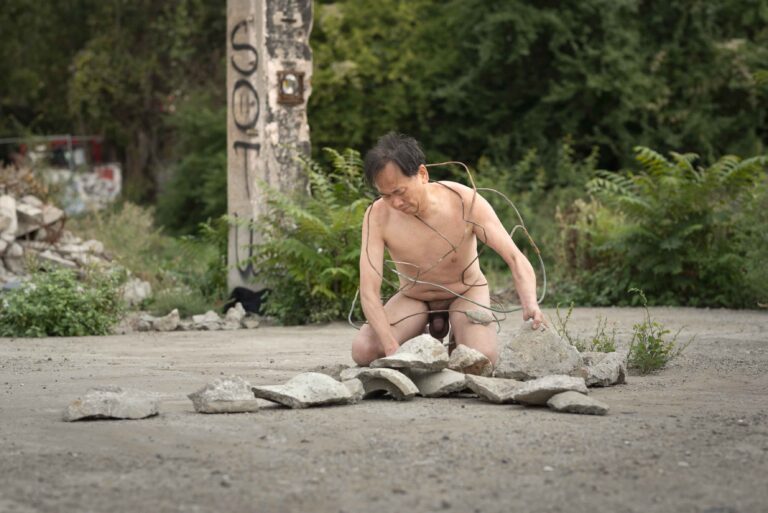 Een naakte man, omgeven door stukken beton en staaldraden, is op zijn knieën aan het werk op een open, verwaarloosde terrein met groene planten op de achtergrond.