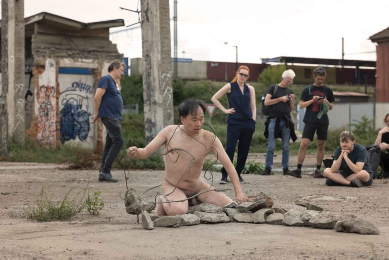 Een naakte performer zit op de grond en is omgeven door een cirkel van stenen, terwijl hij een draad vasthoudt. Op de achtergrond kijken verschillende mensen toe, variërend in hun houding en uitdrukking, met graffiti op de muren achter hen en groen onkruid in de omgeving.