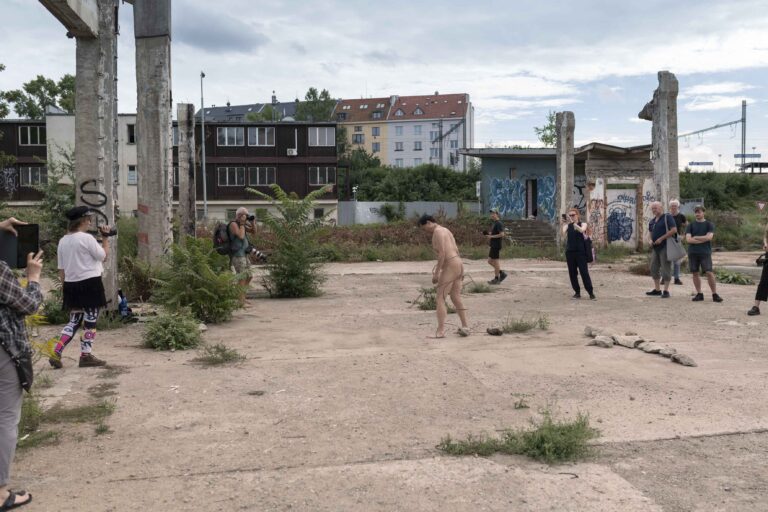 Een naakte man staat in een verlaten gebied tussen verwaarloosde gebouwen met grafitti. Een groep mensen kijkt naar hem en fotografeert de situatie, terwijl er kruiden en planten in de grond groeien.