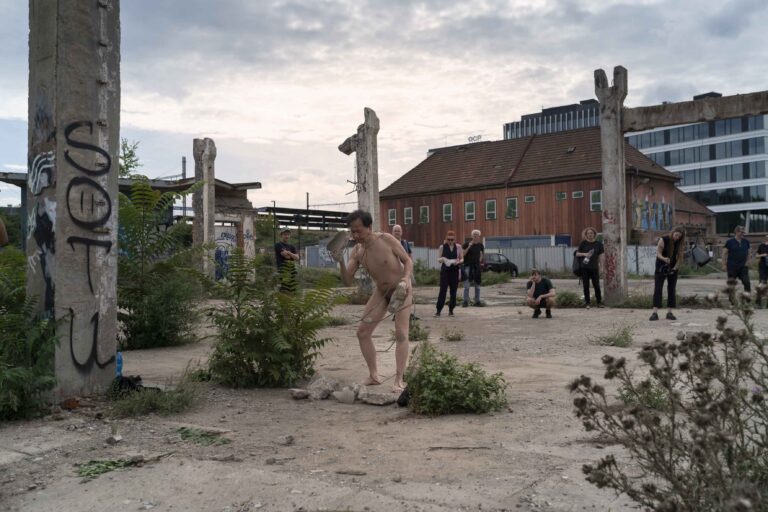 Een naakte persoon staat in een verwoeste omgeving met betonnen pilaren en een hoop puin op de achtergrond; er zijn ook enkele gras- en plantenborders zichtbaar.