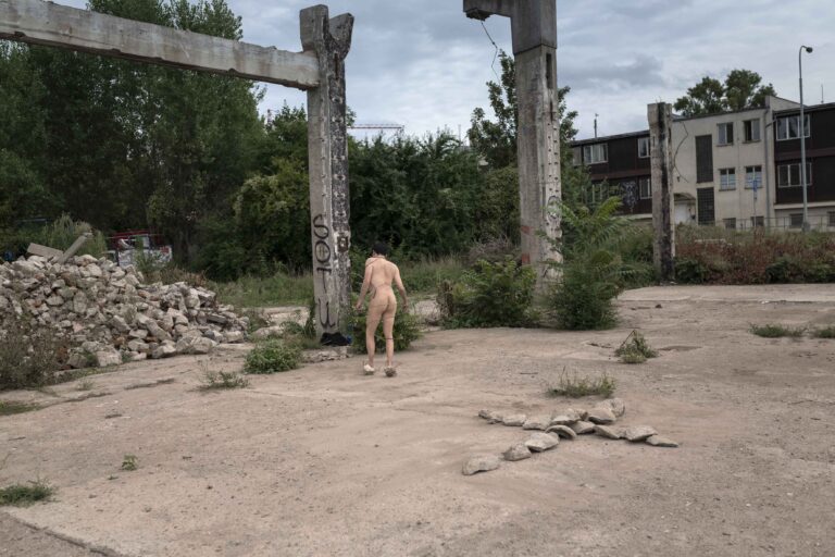 Een naakte persoon staat in een verwoeste omgeving met betonnen pilaren en een hoop puin op de achtergrond; er zijn ook enkele gras- en plantenborders zichtbaar.