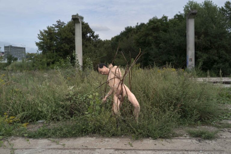 Een naakte persoon staat in een verwilderd gebied vol gras en planten, met takken om hun lichaam gewikkeld, in de achtergrond zijn groene bomen en een gebouw zichtbaar onder een bewolkte lucht.