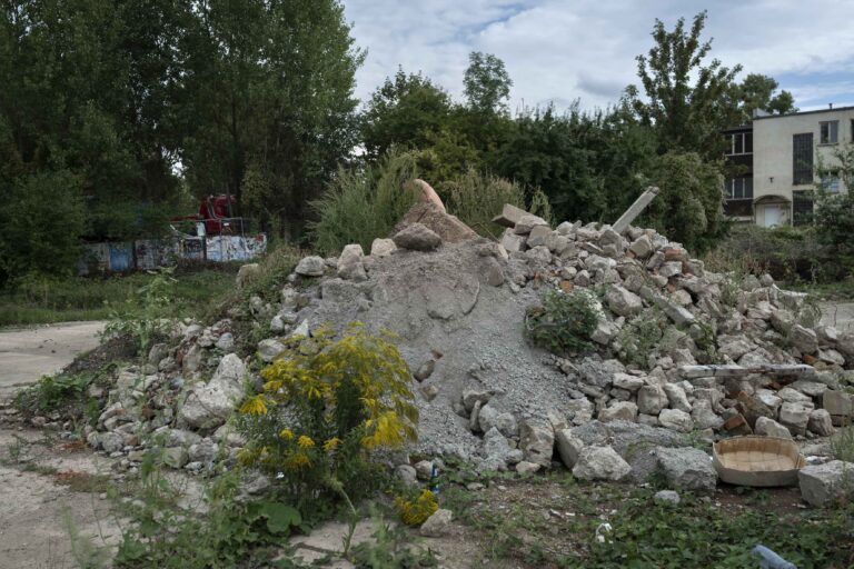 Een hopen puin van stenen en betonstenen op een verlaten terrein, omringd door begroeiing en bomen, met een gedeeltelijk zichtbare, verwaarloosde gebouw op de achtergrond.