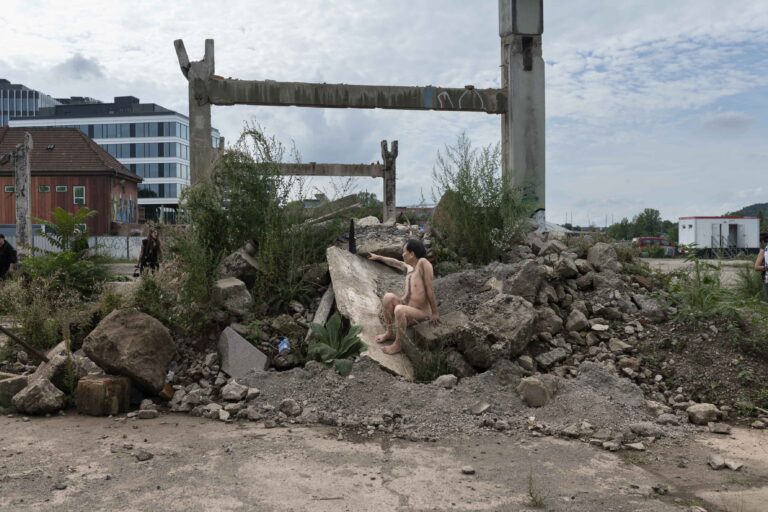 Een naakte persoon zit op een grote betonnen plaat te midden van een puinveld, omringd door stenen, asfalt en begroeiing, met een moderne gebouwen in de achtergrond en enkele personen die voorbijlopen.
