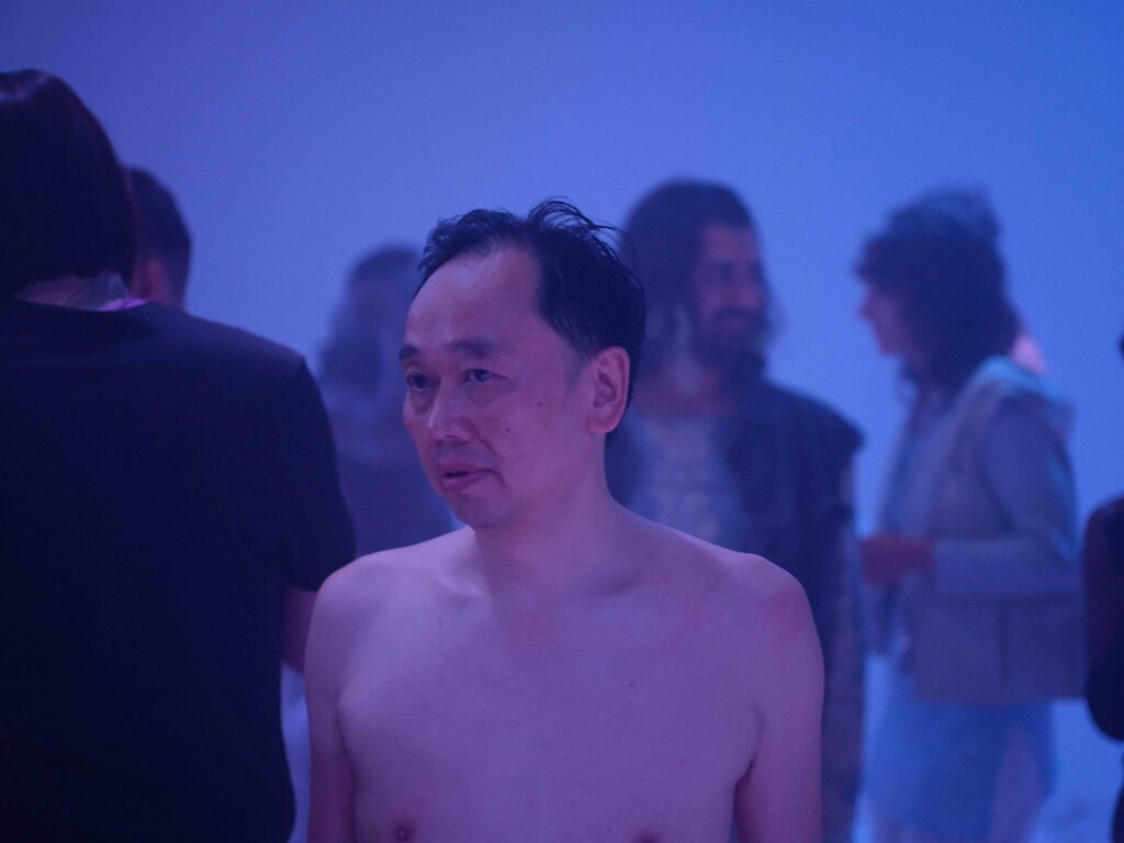 Dirt 3 Een naakte man met kort, donker haar en een neutrale gezichtsuitdrukking staat in een mistige omgeving met een gedimde, blauwe achtergrond. Mensen in de achtergrond zijn vaag zichtbaar en lijken zich te vermaken in de ruimte.