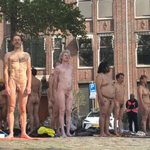 Een groep mensen staat naakt op straat, omgeven door bomen en een bakstenen gebouw; enkele hebben tattoos en er liggen handdoeken op de grond.