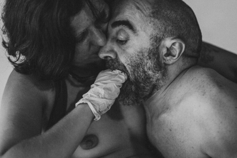 Een groep naakte mensen in een zwart-wit setting, gekleed in verschillende stijlen, die interactie hebben en in verschillende posities zijn geplaatst op een vloer met een heldere achtergrond, met enkele figuren die hun gezichten bedekken en anderen die elkaar omarmen.