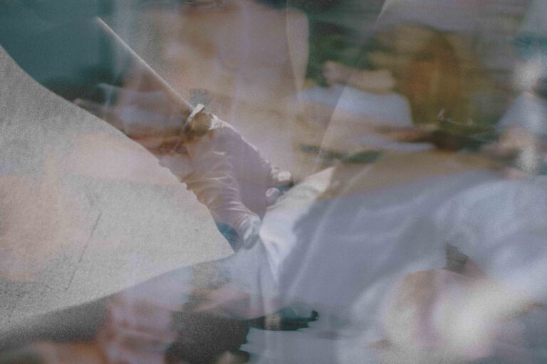 Een wazige afbeelding die de arm van een persoon toont, gekleed in een witte langemouwige kleding, terwijl ze iets vasthouden met een vage achtergrond van andere mensen en objecten.