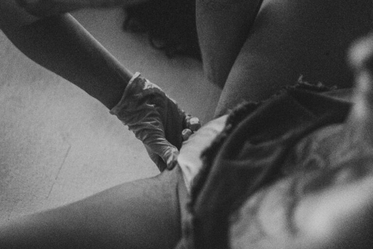 Een groep naakte mensen in een zwart-wit setting, gekleed in verschillende stijlen, die interactie hebben en in verschillende posities zijn geplaatst op een vloer met een heldere achtergrond, met enkele figuren die hun gezichten bedekken en anderen die elkaar omarmen.