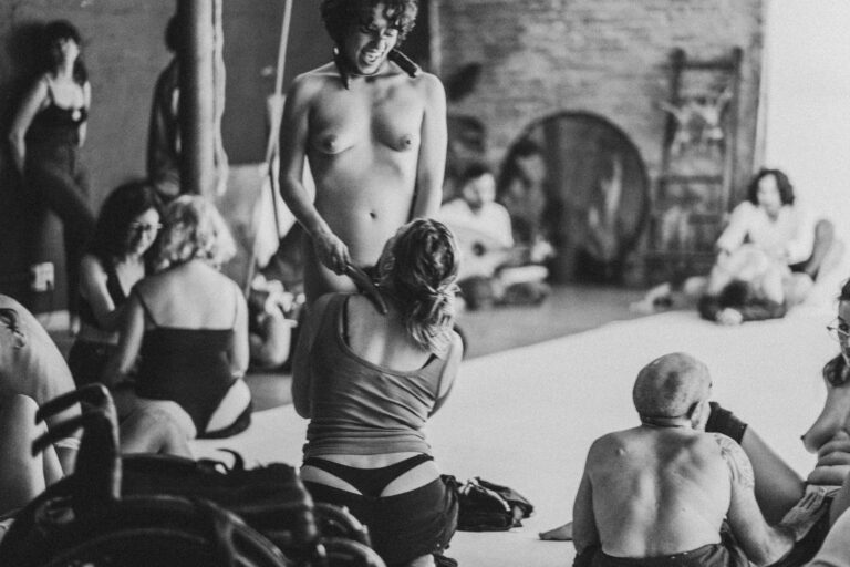 Een groep naakte mensen in een zwart-wit setting, gekleed in verschillende stijlen, die interactie hebben en in verschillende posities zijn geplaatst op een vloer met een heldere achtergrond, met enkele figuren die hun gezichten bedekken en anderen die elkaar omarmen.