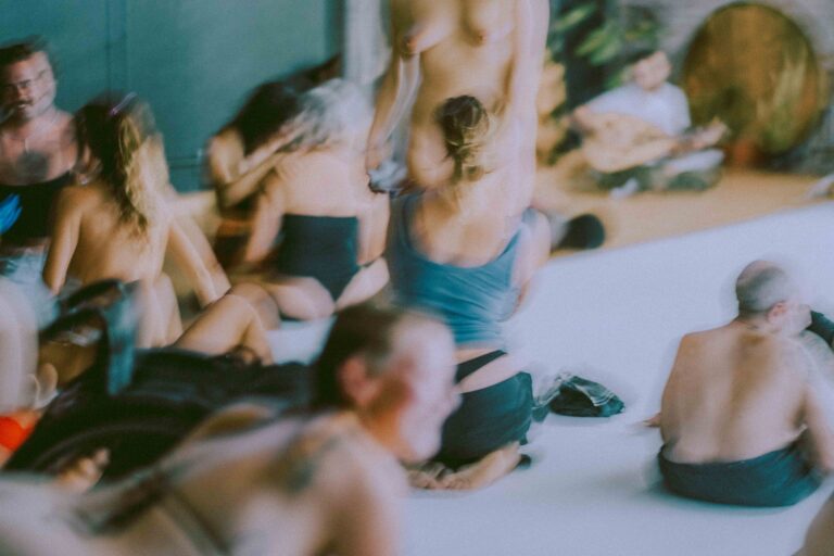 Een groep mensen die zich bezig houden met lichaamswerk of groepsinteractie, met een mix van naakte en geklede individuen die elkaar steun bieden en hun gevoelens delen in een sombere omgeving.