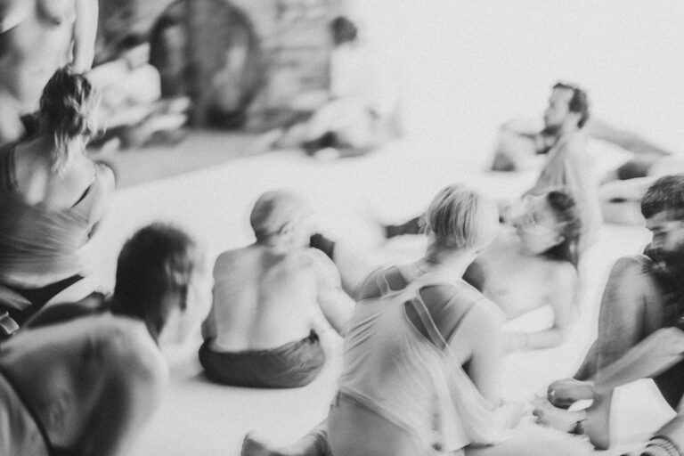 Een groep naakte mensen in een zwart-wit setting, gekleed in verschillende stijlen, die interactie hebben en in verschillende posities zijn geplaatst op een vloer met een heldere achtergrond, met enkele figuren die hun gezichten bedekken en anderen die elkaar omarmen.