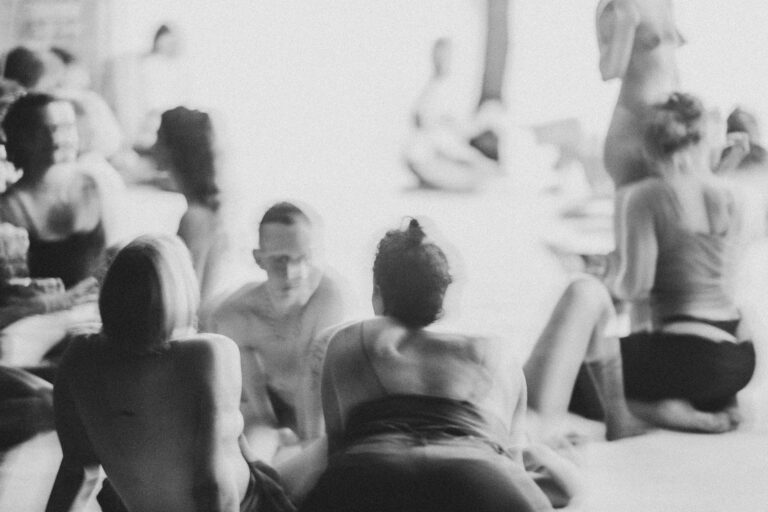 Een groep naakte mensen in een zwart-wit setting, gekleed in verschillende stijlen, die interactie hebben en in verschillende posities zijn geplaatst op een vloer met een heldere achtergrond, met enkele figuren die hun gezichten bedekken en anderen die elkaar omarmen.