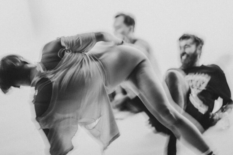 Een groep naakte mensen in een zwart-wit setting, gekleed in verschillende stijlen, die interactie hebben en in verschillende posities zijn geplaatst op een vloer met een heldere achtergrond, met enkele figuren die hun gezichten bedekken en anderen die elkaar omarmen.