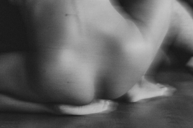 Een abstracte zwart-wit afbeelding van een naaktfiguur die zich op de grond bevindt, met focus op de rondingen van de rug en de voeten, die in een ontspannen positie zijn geplaatst.