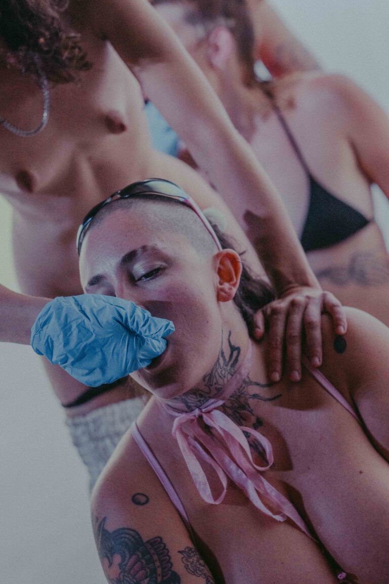 Multiple nude figures in intimate contact with colorful aesthetic, showing bare torsos with tattoos, captured with vibrant pink and blue tones.