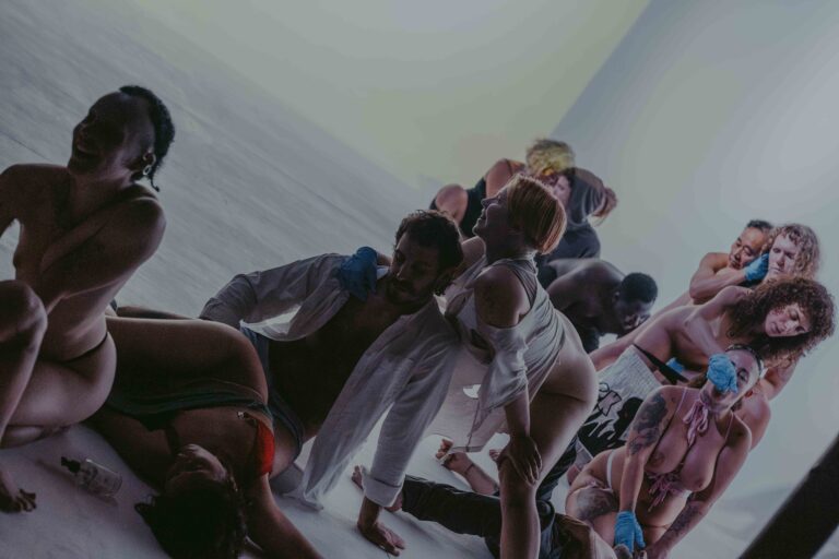 Een groep mensen die zich bezig houden met lichaamswerk of groepsinteractie, met een mix van naakte en geklede individuen die elkaar steun bieden en hun gevoelens delen in een sombere omgeving.