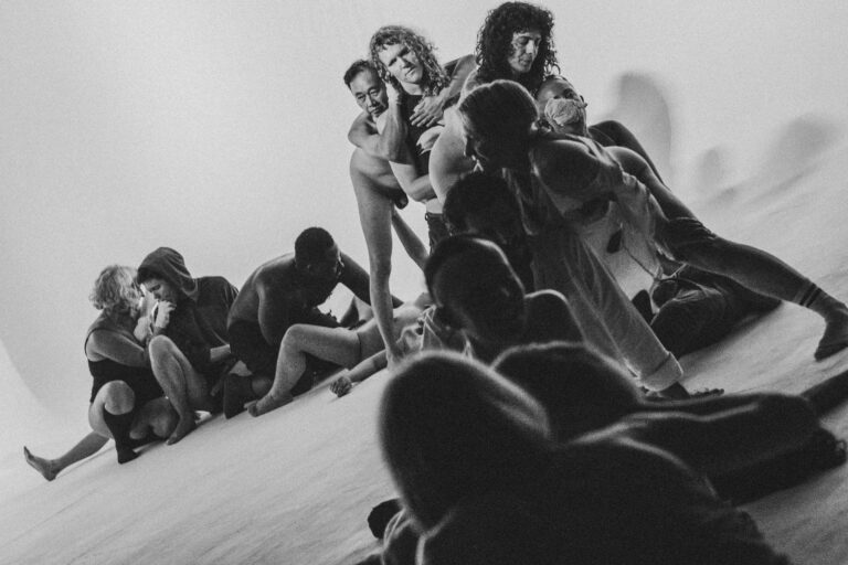 Een groep naakte mensen in een zwart-wit setting, gekleed in verschillende stijlen, die interactie hebben en in verschillende posities zijn geplaatst op een vloer met een heldere achtergrond, met enkele figuren die hun gezichten bedekken en anderen die elkaar omarmen.