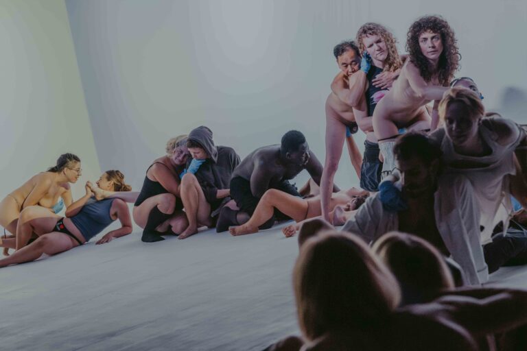 Een groep mensen die zich bezig houden met lichaamswerk of groepsinteractie, met een mix van naakte en geklede individuen die elkaar steun bieden en hun gevoelens delen in een sombere omgeving.