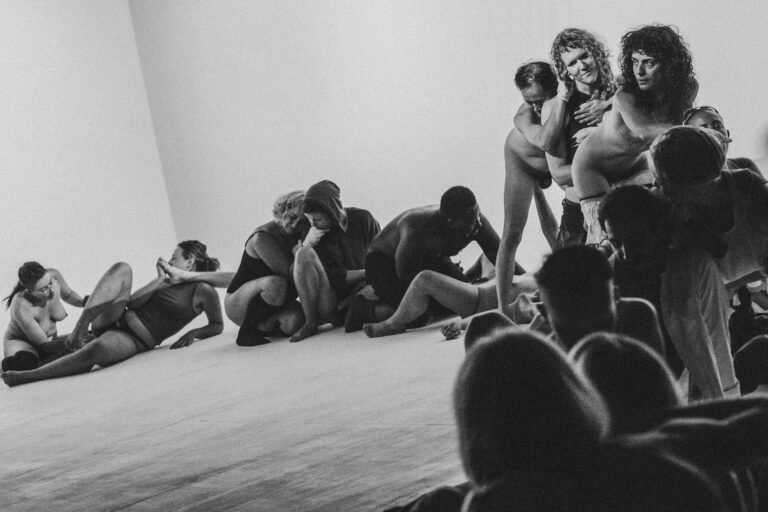 Een groep mensen in een artistieke scène, elkaar omarmend en op de grond zittend, met een neutrale achtergrond en in zwart-wit; de focus ligt op hun interacties en poses.