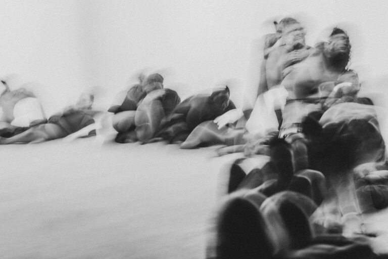 Een abstracte, onscherpe afbeelding van een groep mensen in een dynamische pose, waarbij ze op de grond liggen en zich in een gezamenlijke beweging lijken te bevinden, met een lichte overbelichting.