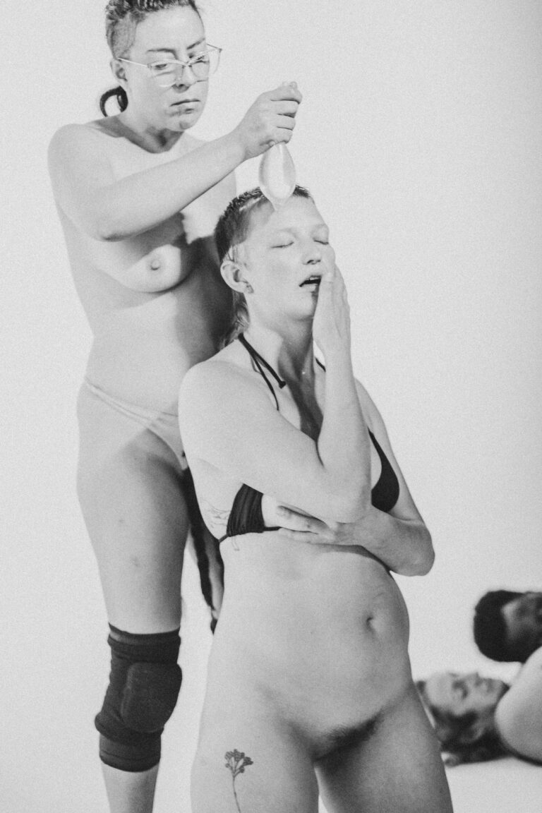 Two women in a black and white photo, one topless and one fully naked placing a flower in hair