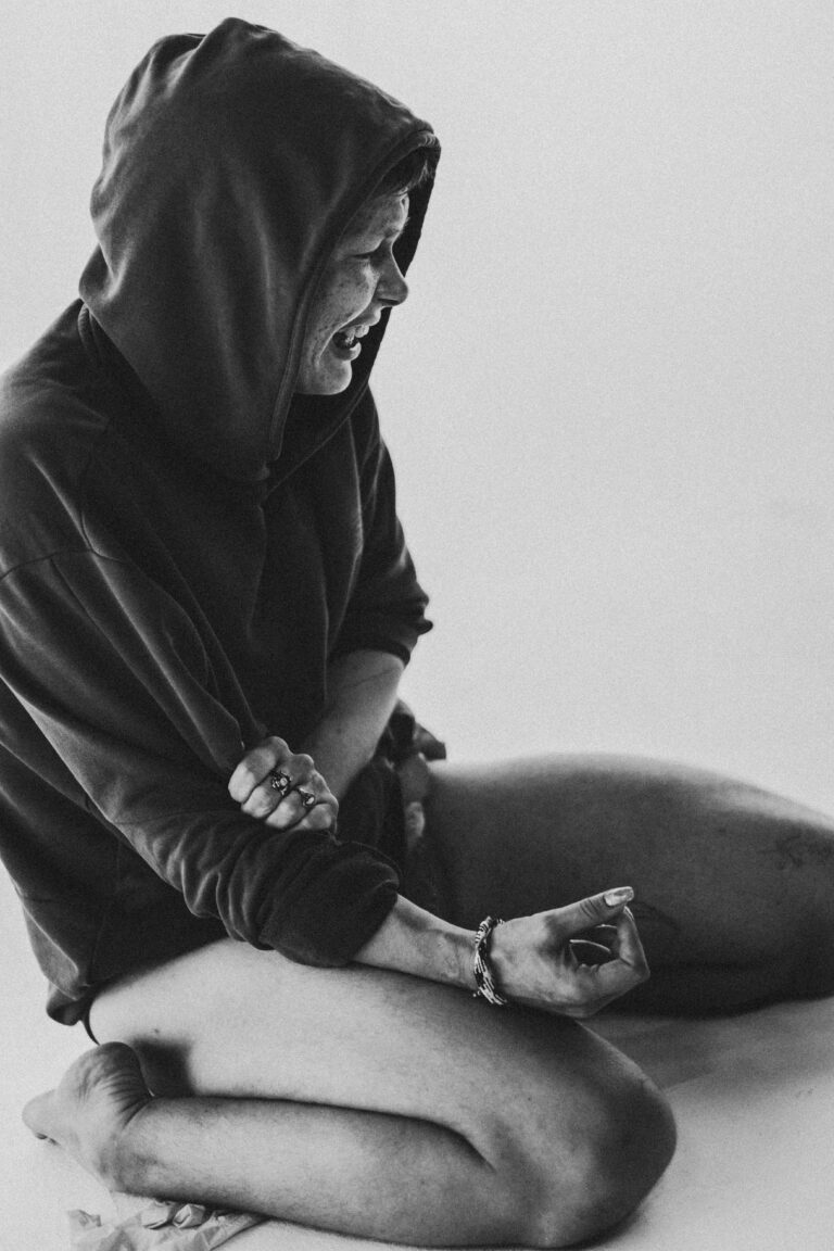 Naked person sitting cross-legged on the ground dressed in a black hoodie with hands in meditation pose