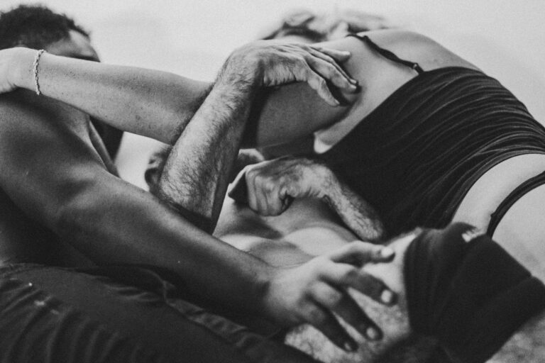 Zwart-wit afbeelding van een groep mensen die een intieme pose aannemen, met focus op hun armen en handen die elkaar vasthouden en omarmen, op een bed of een zachte ondergrond.