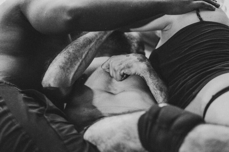 Drie mensen liggen op de vloer, omringd door elkaar, met een focus op de handen en armen die verschillende posities aannemen, waarbij een hand met een tatoeage zichtbaar is. De afbeelding is in zwart-wit met een intieme sfeer.