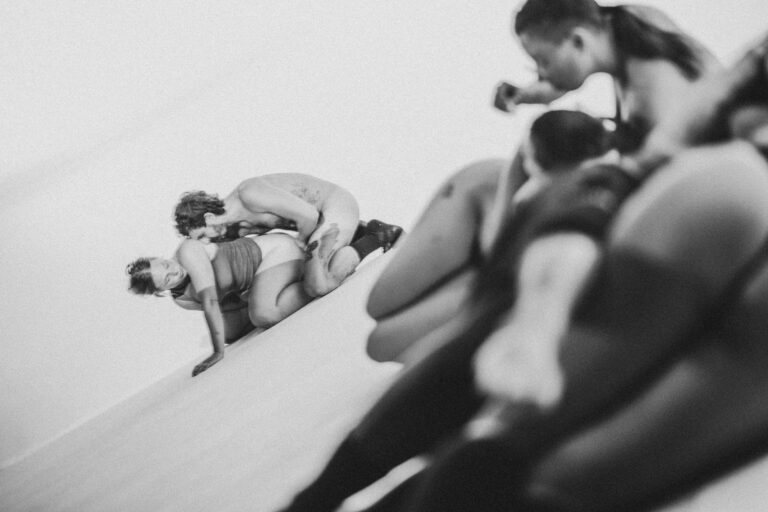 Een groep naakte mensen met Philemon Mukarno die elkaar ondersteunen in een acrobatische houding, met een persoon in het midden die door anderen wordt vastgehouden. De foto is in zwart-wit en geeft een artistieke sfeer weer, met een focus op de menselijke vormen en bewegingen.