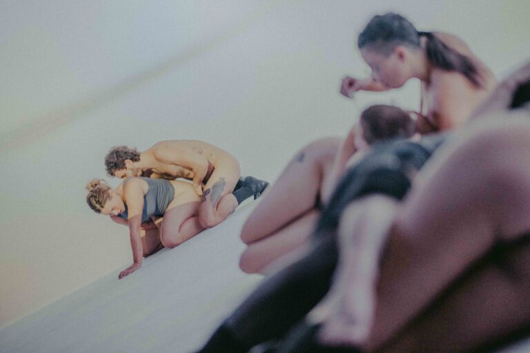 Een groep mensen die zich bezig houden met lichaamswerk of groepsinteractie, met een mix van naakte en geklede individuen die elkaar steun bieden en hun gevoelens delen in een sombere omgeving.