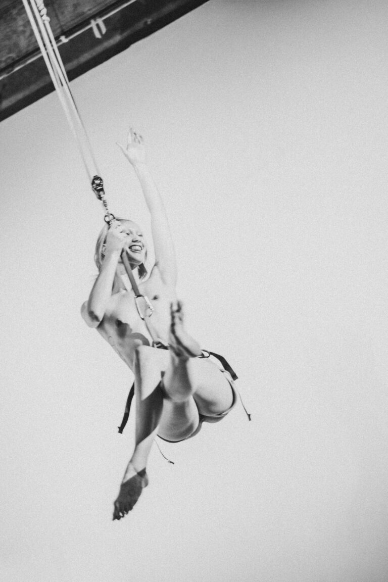 Naked person performing an acrobatic aerial movement on chains in mid-air with legs spread