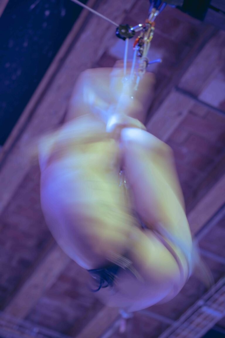 A nude acrobat hanging upside down from a rope with heavy motion blur, captured in purple and blue tones with ethereal lighting.