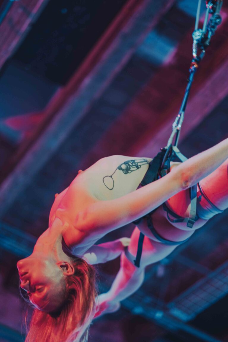 A nude acrobat hanging upside down from a rope with bare legs extended, captured in vibrant pink, red and blue lighting.
