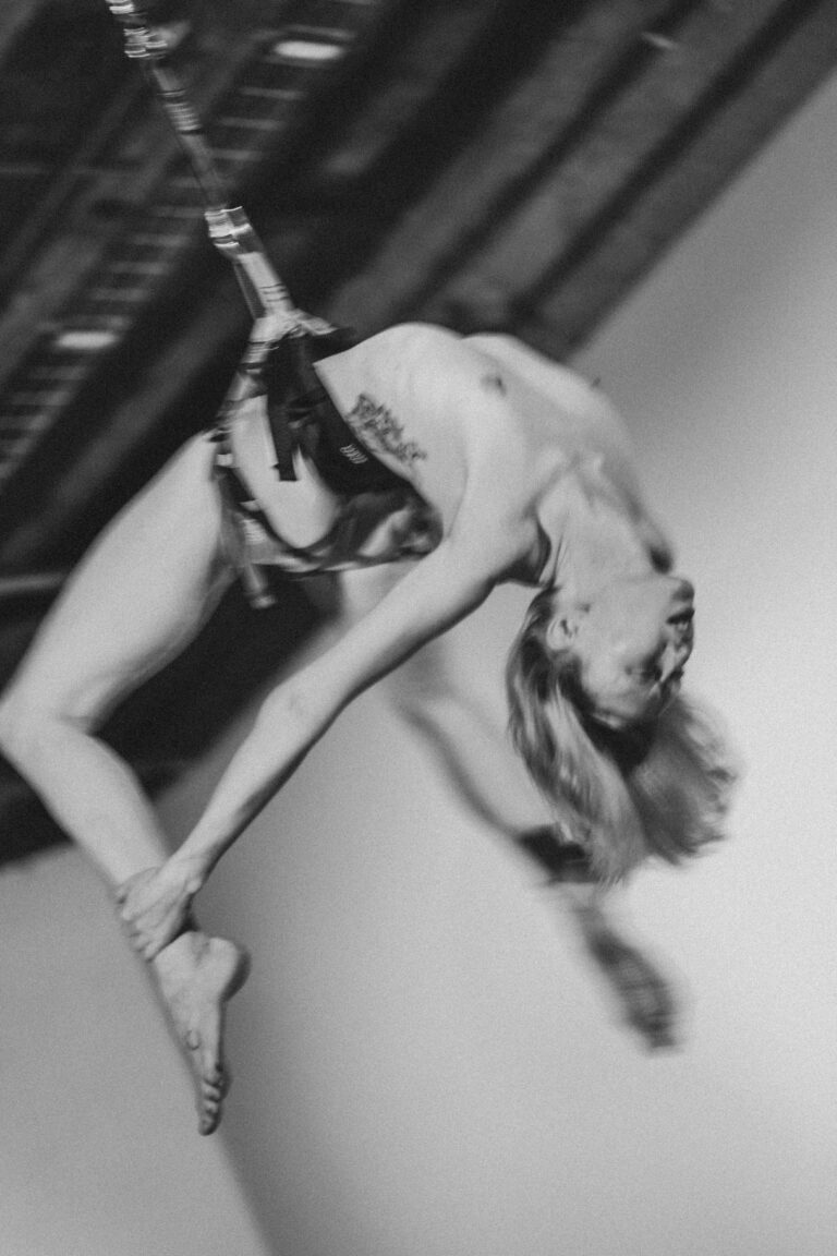 Naked person hanging from a rope in an acrobatic aerial pose with head tilted back