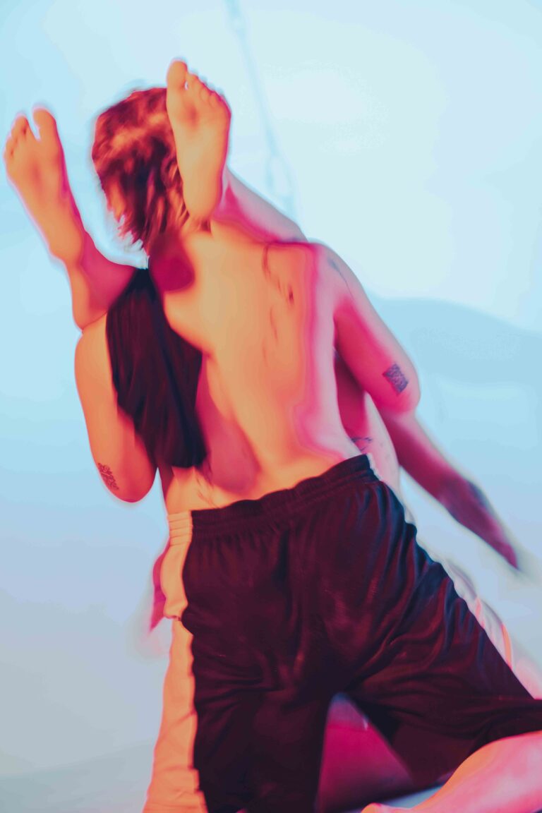 Two people in a dynamic pose with their arms raised, one shirtless and one bare-backed, captured in vibrant pink and orange tones against a soft blue background.