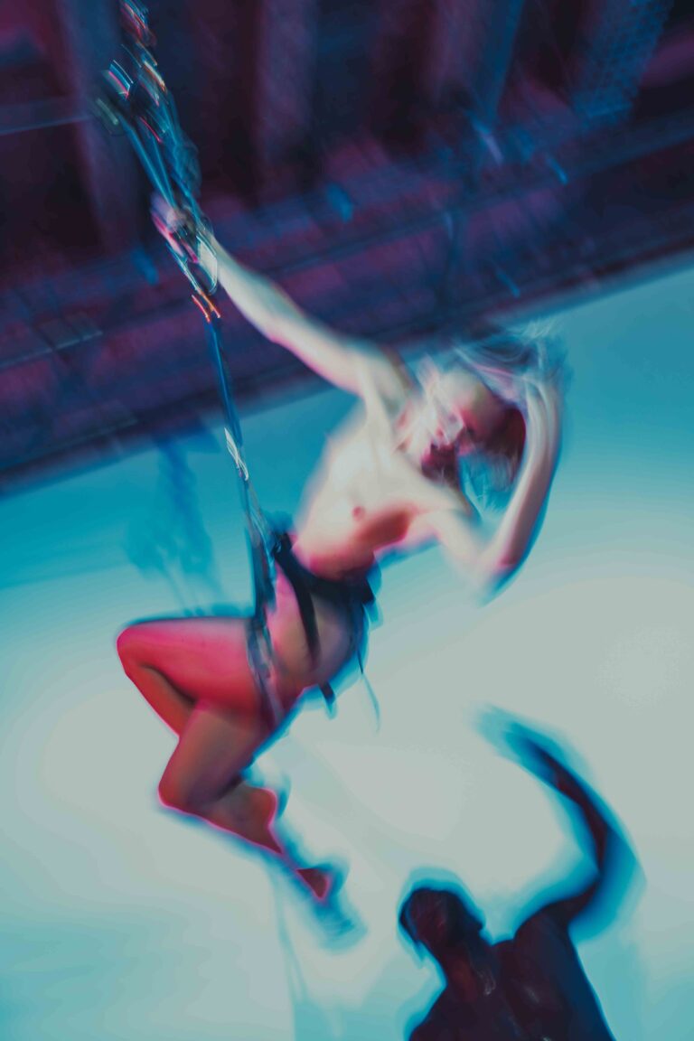 A nude performer suspended in mid-air, supported by another performer below, captured in dramatic blue and pink lighting that creates a dynamic aerial performance scene.
